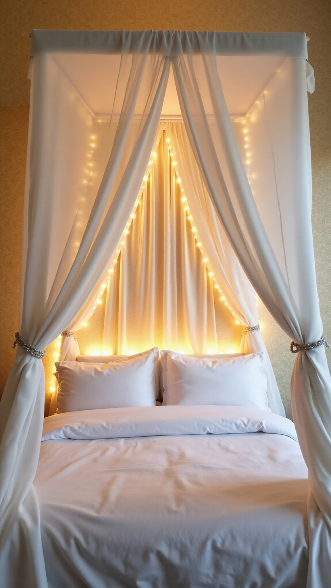 Twin bed with sheer white canopy and warm LED lighting in a small, gold-accented bedroom at sunset.