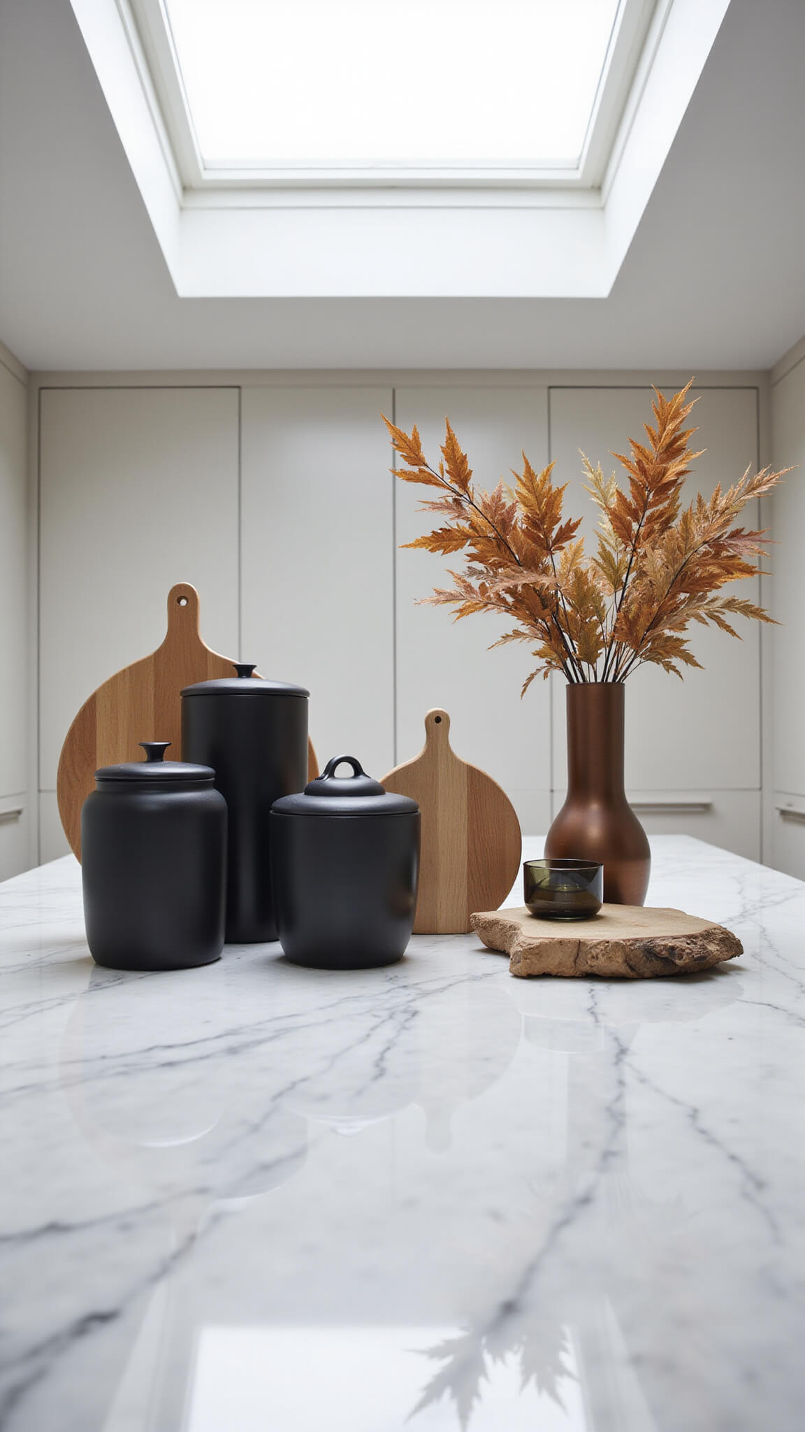 Overhead view of modern kitchen vignette with 6ft marble island, matte black accessories in groups of three, bronze vase with dried oak leaves, lit by skylight and side fill light.