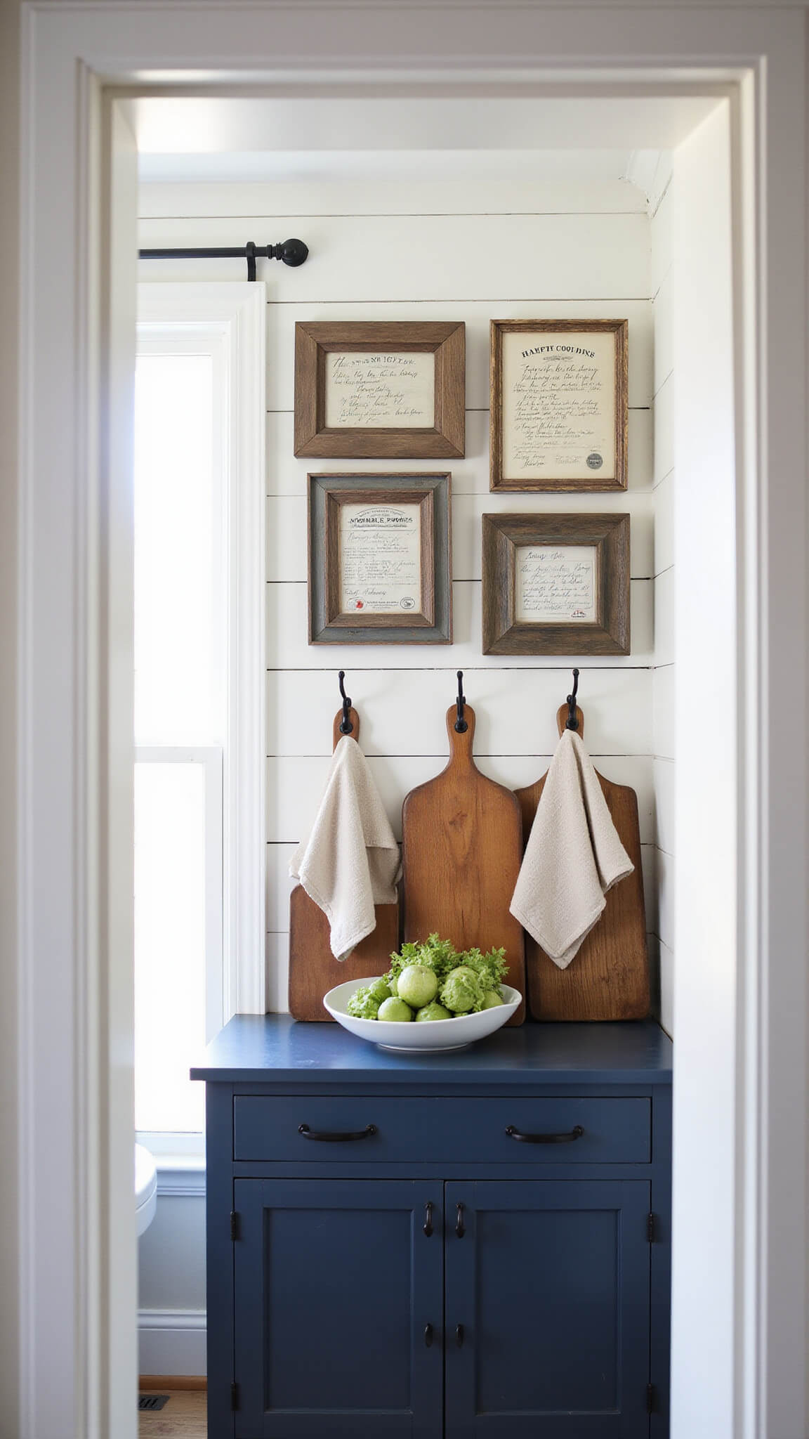 Morning light filters through sheer curtains into a cozy farmhouse kitchen with vintage cooking signs, framed recipe cards above a navy blue sideboard, warm wood cutting boards, and linen tea towels on iron hooks.