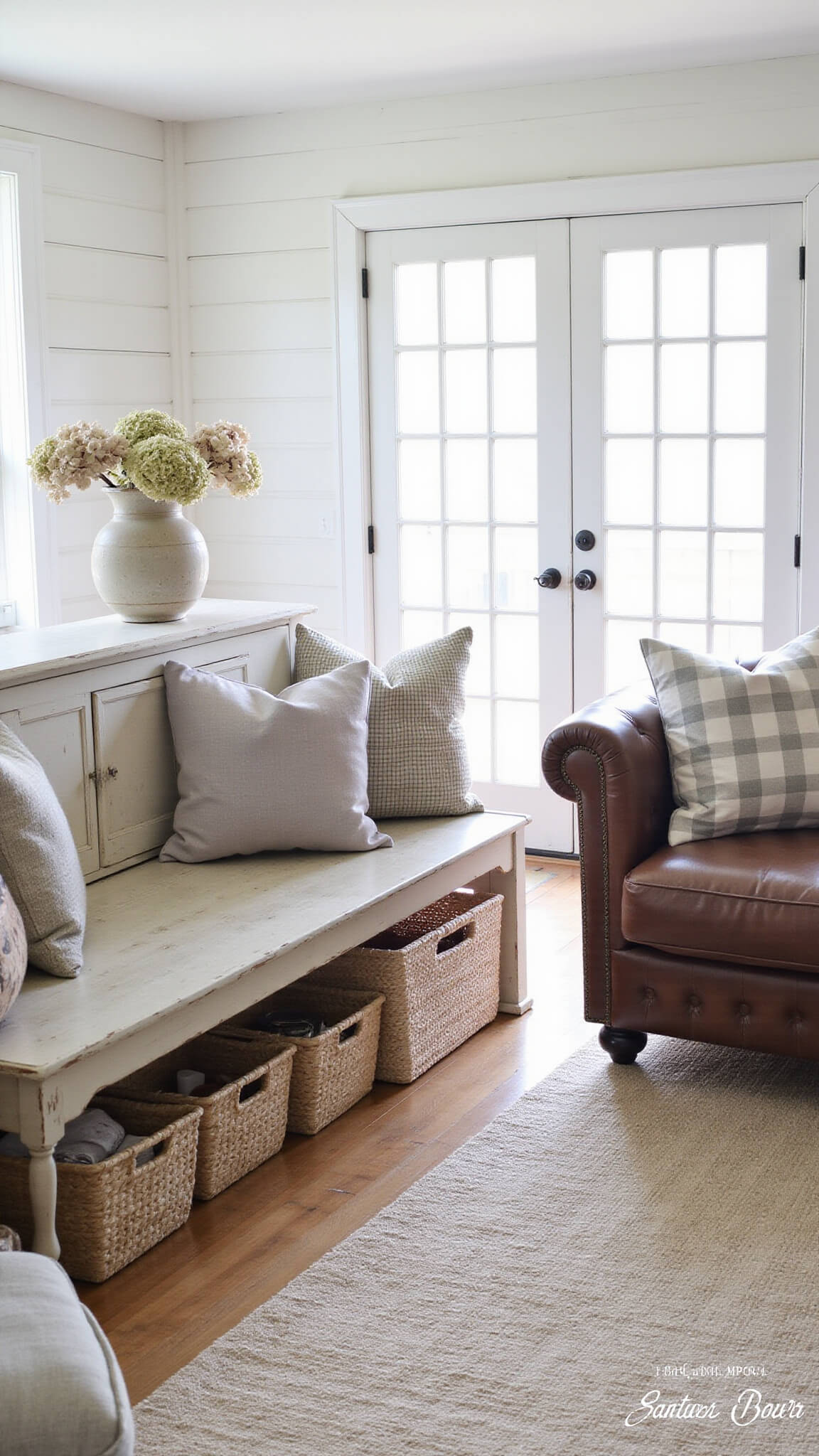 Farmhouse living room with vintage furnishings bathed in morning light from east-facing French doors, featuring a distressed leather Chesterfield sofa, textured pillows, and dried hydrangeas against white shiplap walls.