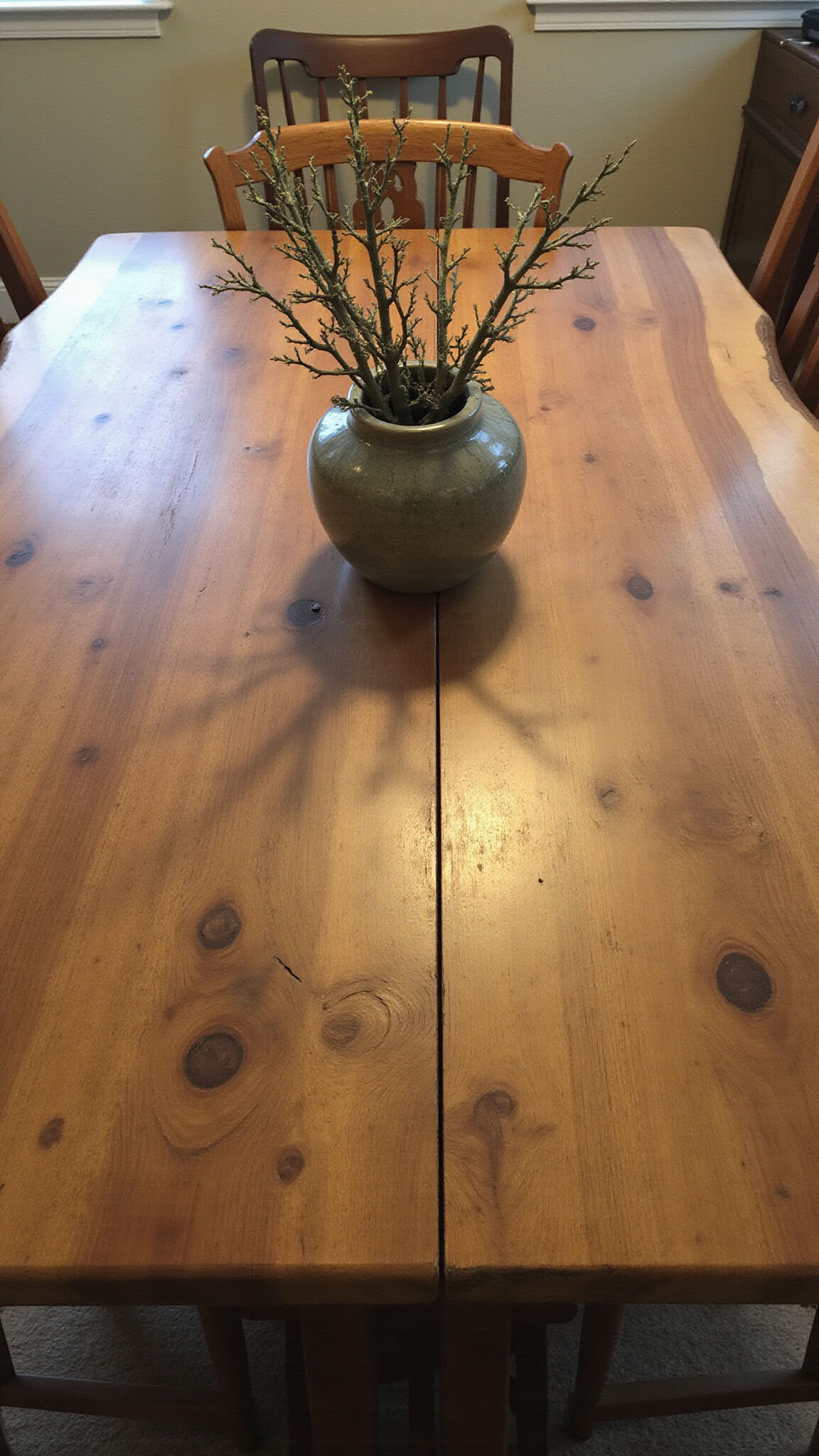 Overhead view of a 10x10ft dining area with a live-edge wooden table featuring natural splits and knots, surrounded by mismatched vintage chairs with aged patinas; a crackle-glazed vessel holding foraged branches sits at center under warm evening light.