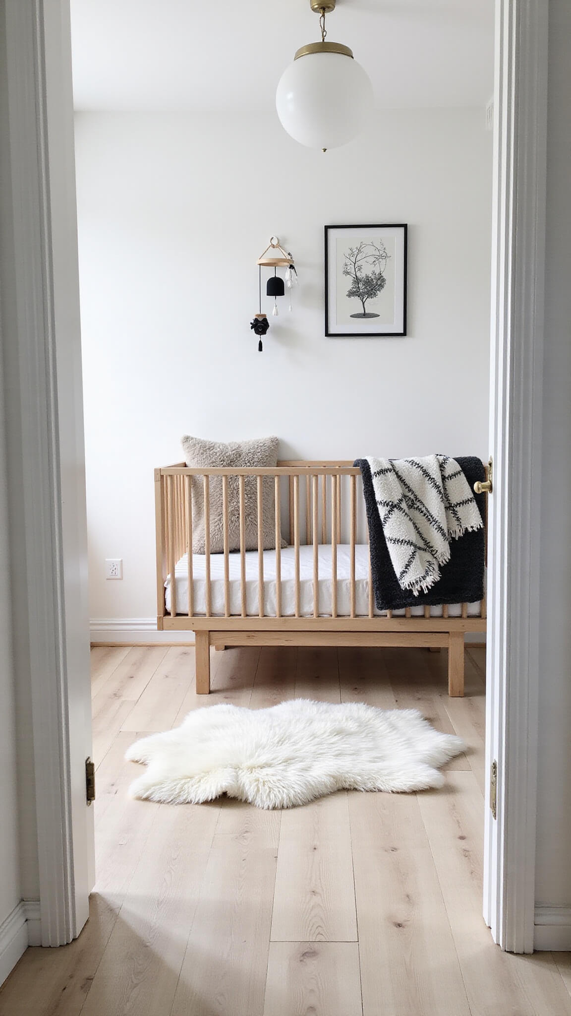 Scandinavian-style nursery with white walls, pale birch floors, wooden crib with monochrome bedding, geometric mobile, and sheepskin throws, bathed in soft early morning light.
