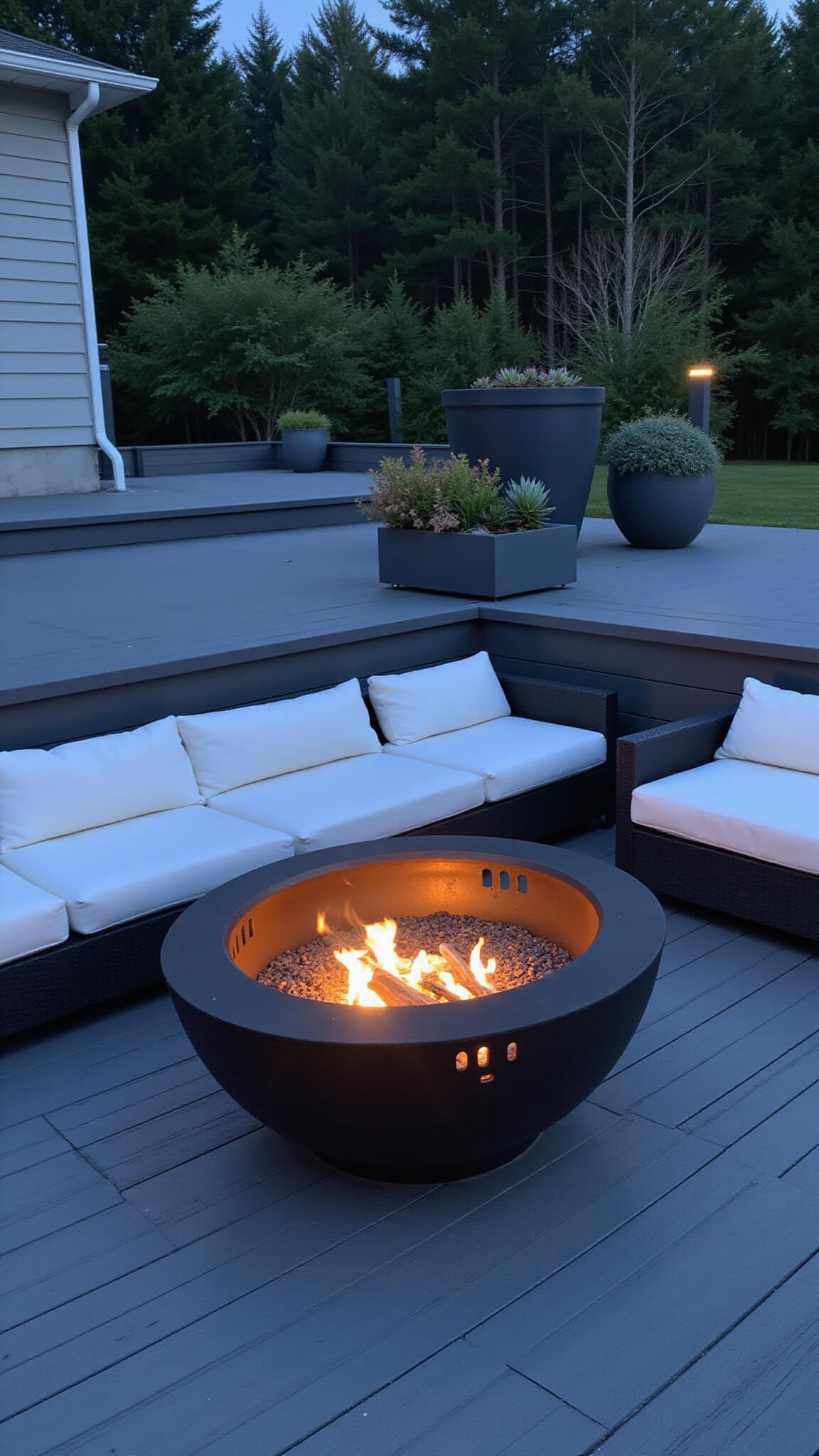 Modern black steel fire bowl with geometric cutouts on composite deck, surrounded by gray sectional seating with white cushions, architectural succulents, and subtle LED lighting at twilight.