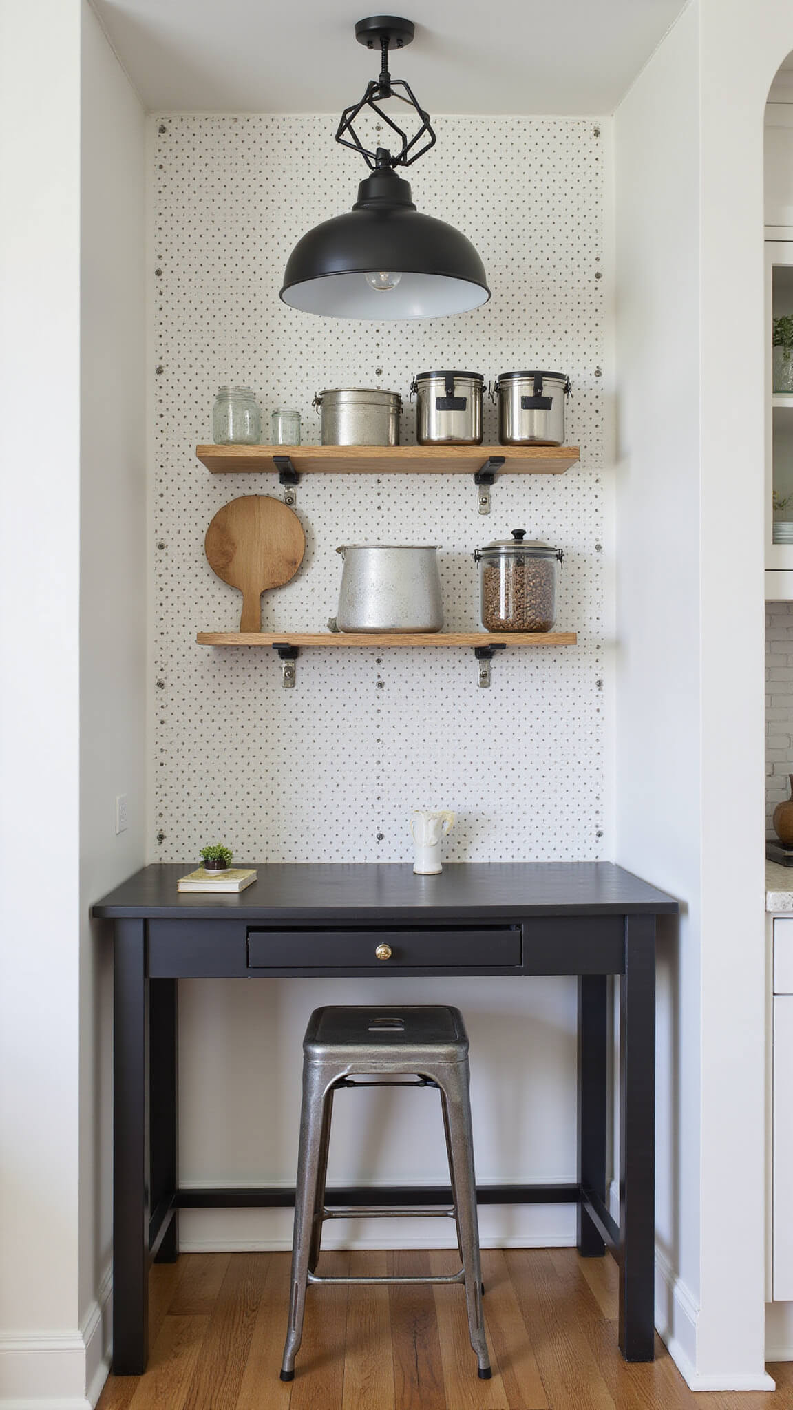 Modern kitchen nook with white pegboard wall, wooden shelves, metal containers, black console desk, industrial stool, and geometric pendant light casting shadows.