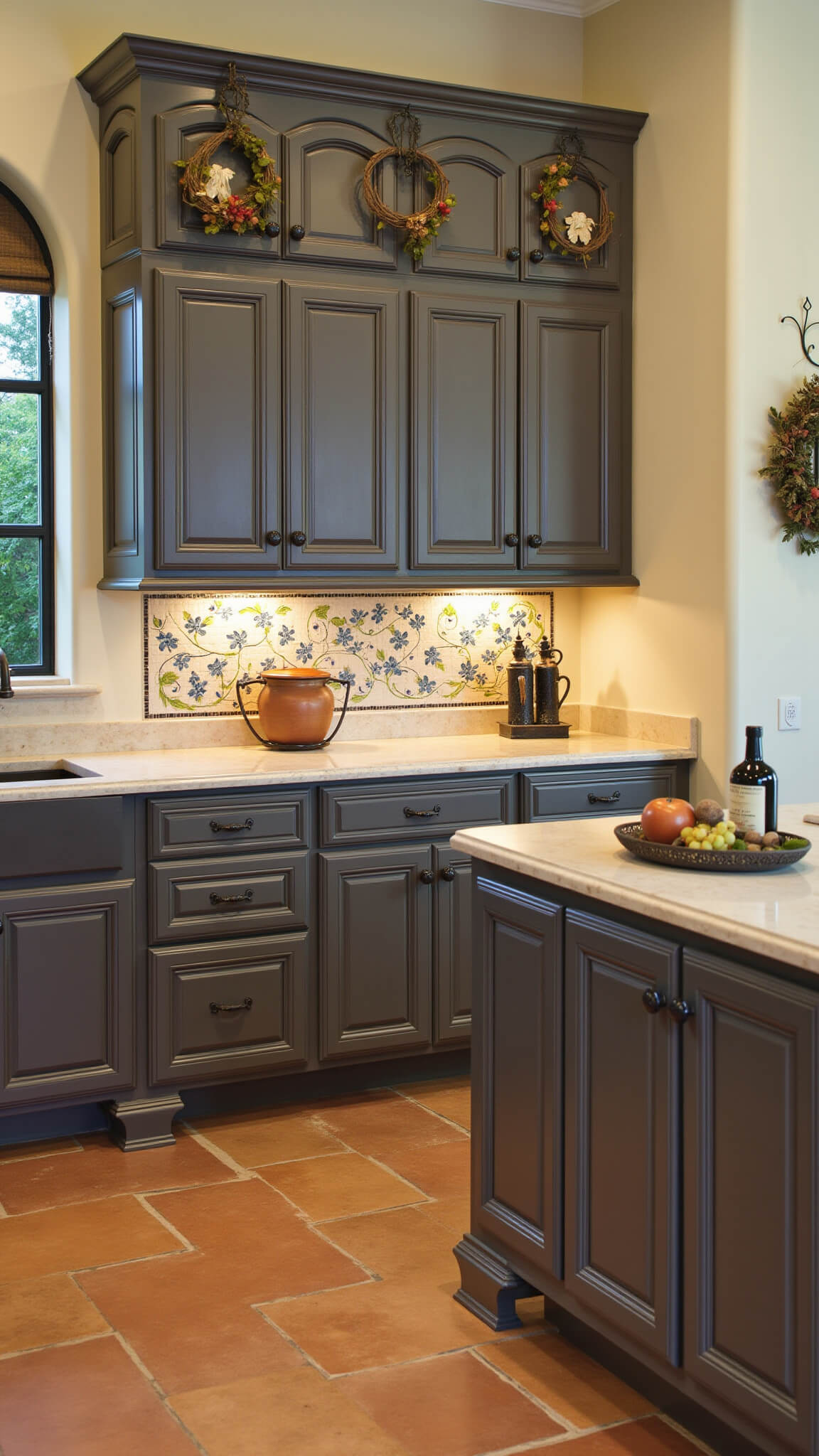 Mediterranean-style 15x17ft kitchen with dark taupe arched cabinets, blue and cream hand-painted tile backsplash, limestone counters, terracotta floors, and copper pots under natural midday light.