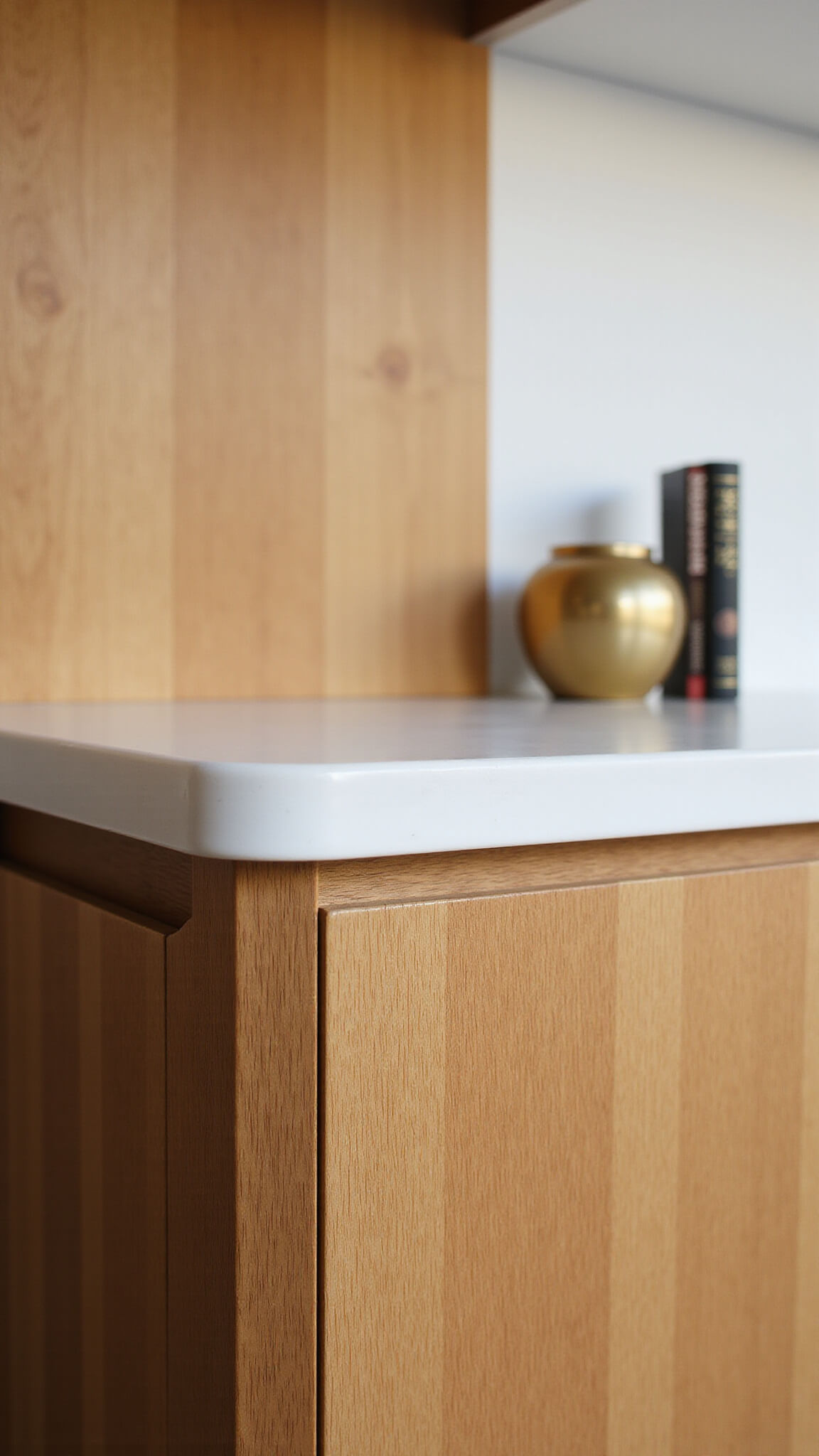 Close-up of oak cabinet corner with white quartz countertop in evening light, featuring brass vessel and cookbook stand.