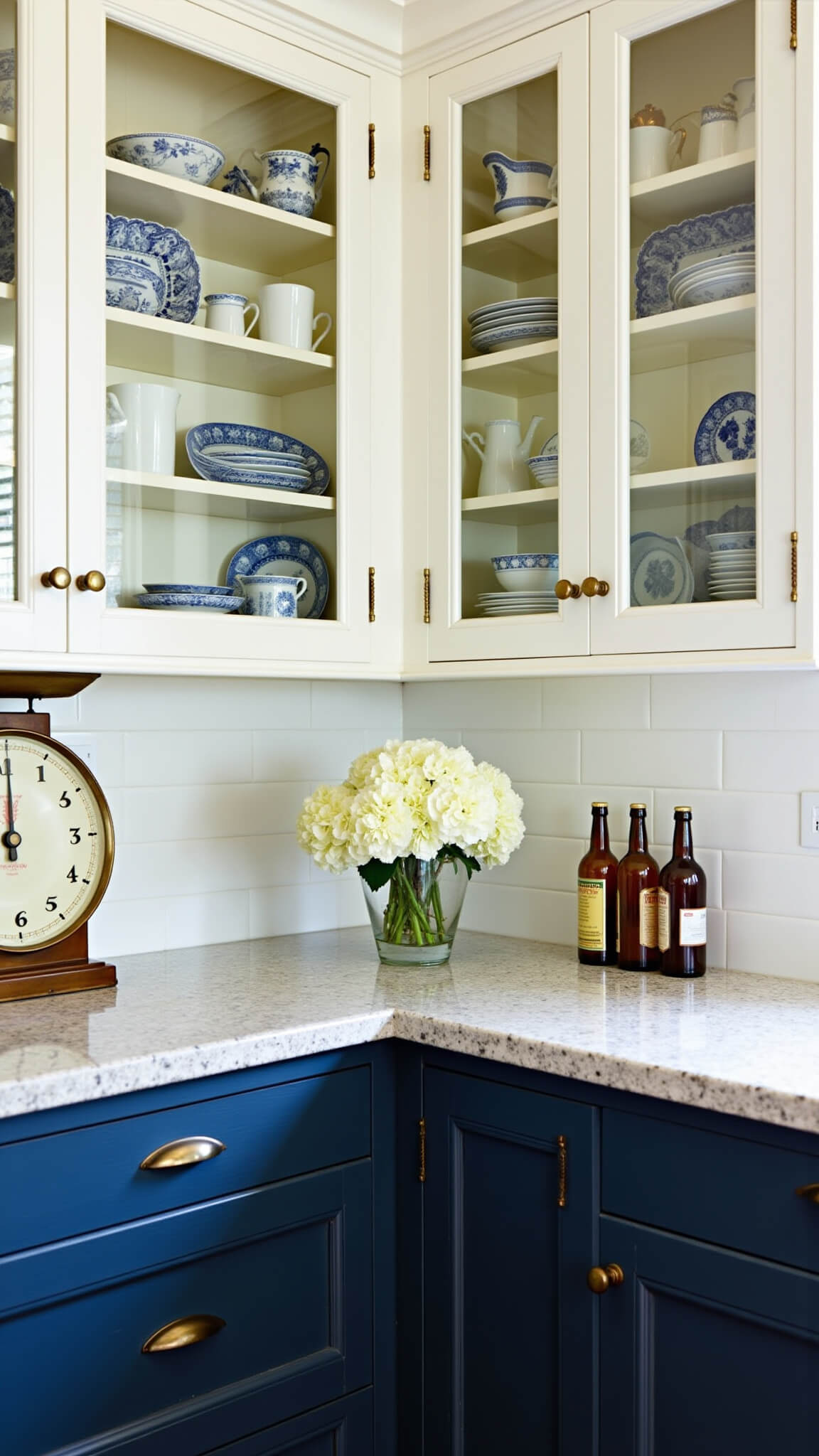 Corner kitchen cabinet display with navy lower cabinets, white glass-front uppers showcasing blue and white china, brass lighting, hydrangeas, antique scale, and amber oil bottles on granite countertop.