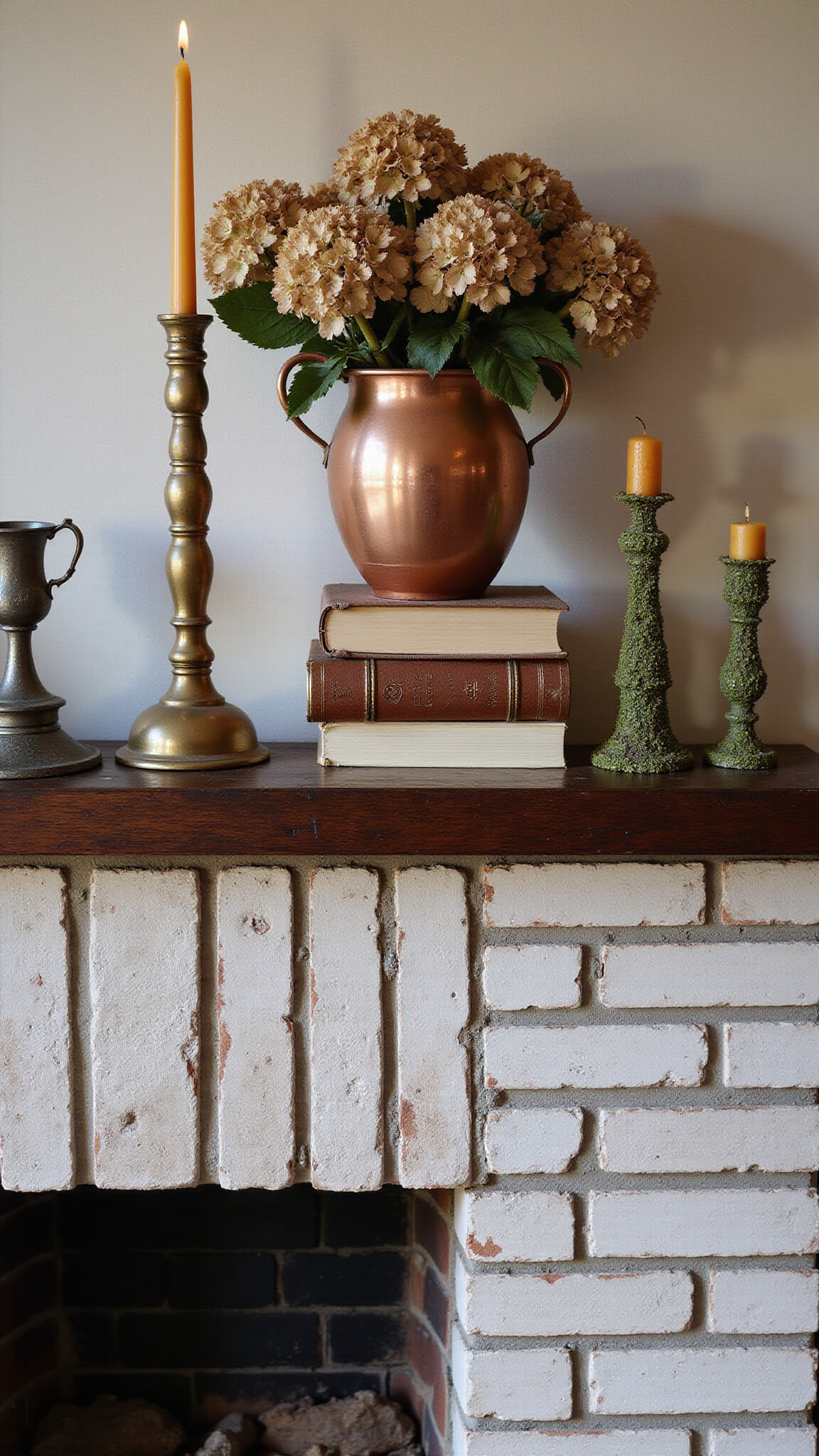 Cozy fireplace vignette with brass tools, dried hydrangeas in copper vase, vintage books, and dripping beeswax candles on textured brick hearth.