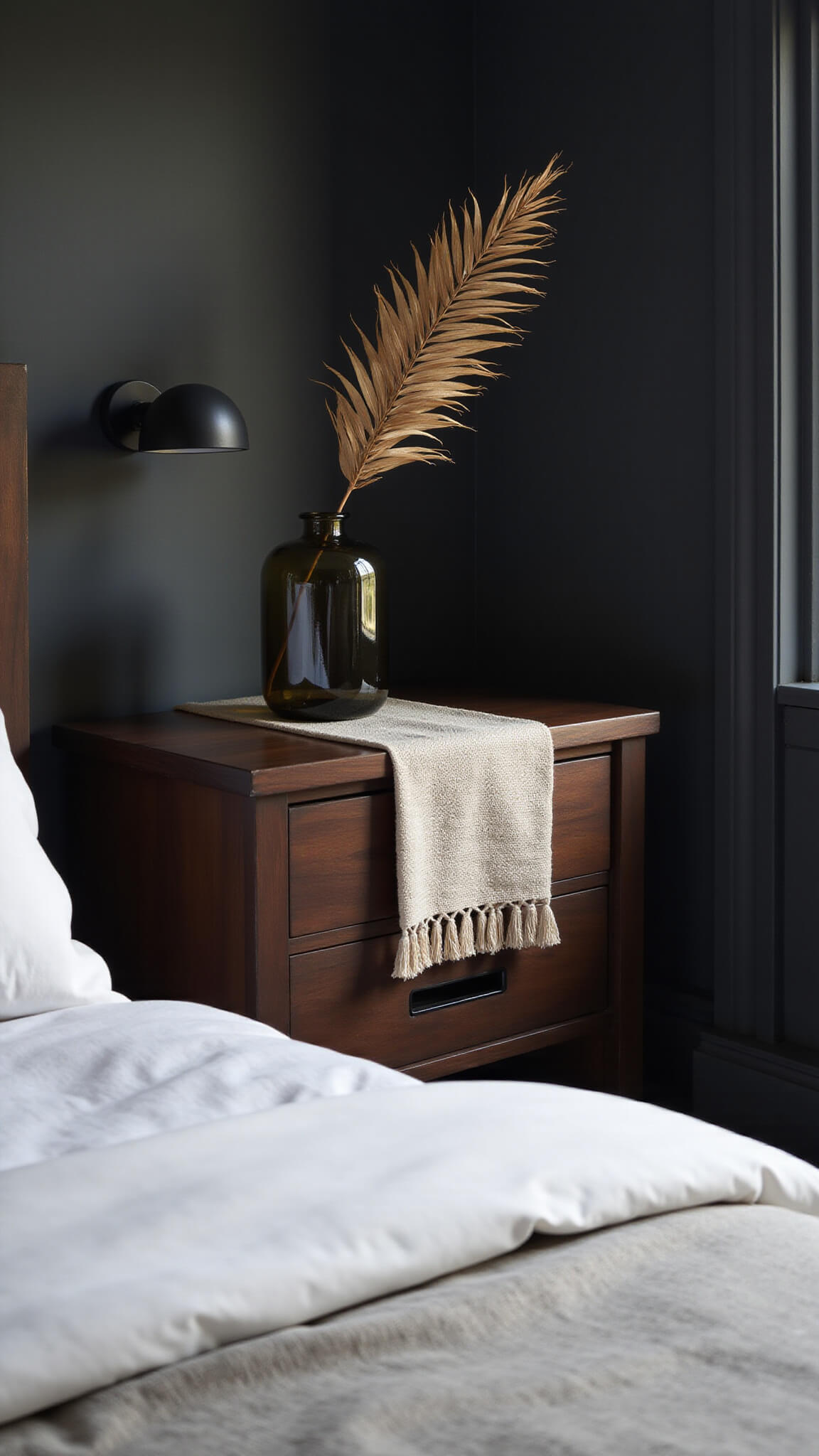 Close-up of black ash Japandi nightstand with architectural drawer pulls, draped linen runner, and smoke glass vase holding dried palm frond, set against deep charcoal walls in soft late afternoon light.