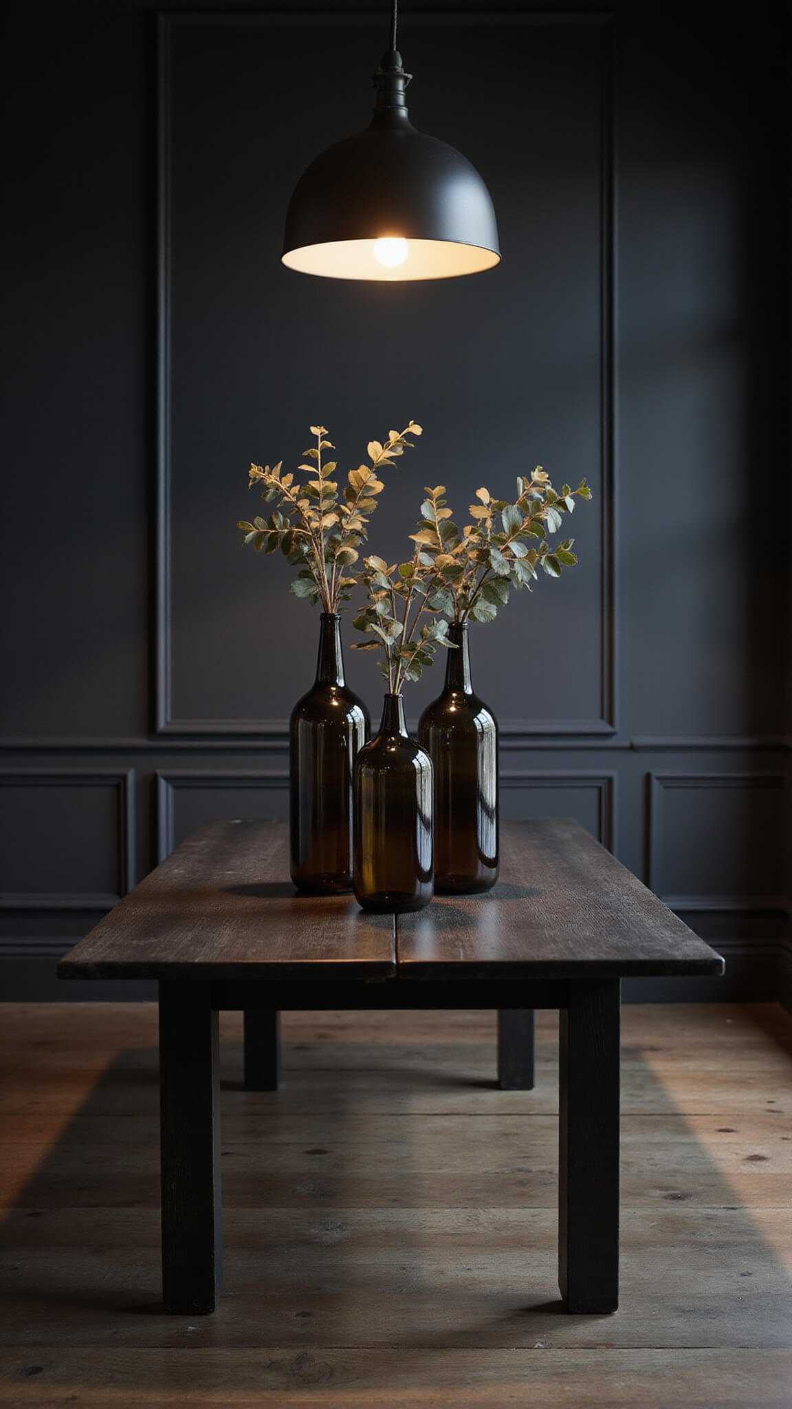 Moody dining room at dusk with a black oak table, three tall smoky glass vases holding dried eucalyptus, dark charcoal walls, and dramatic side lighting.
