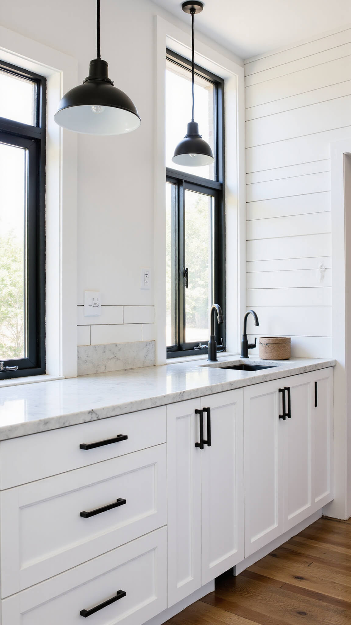Modern farmhouse kitchen with white flat-panel cabinets, black hardware, marble island, and industrial pendant lights, viewed from elevated corner angle.