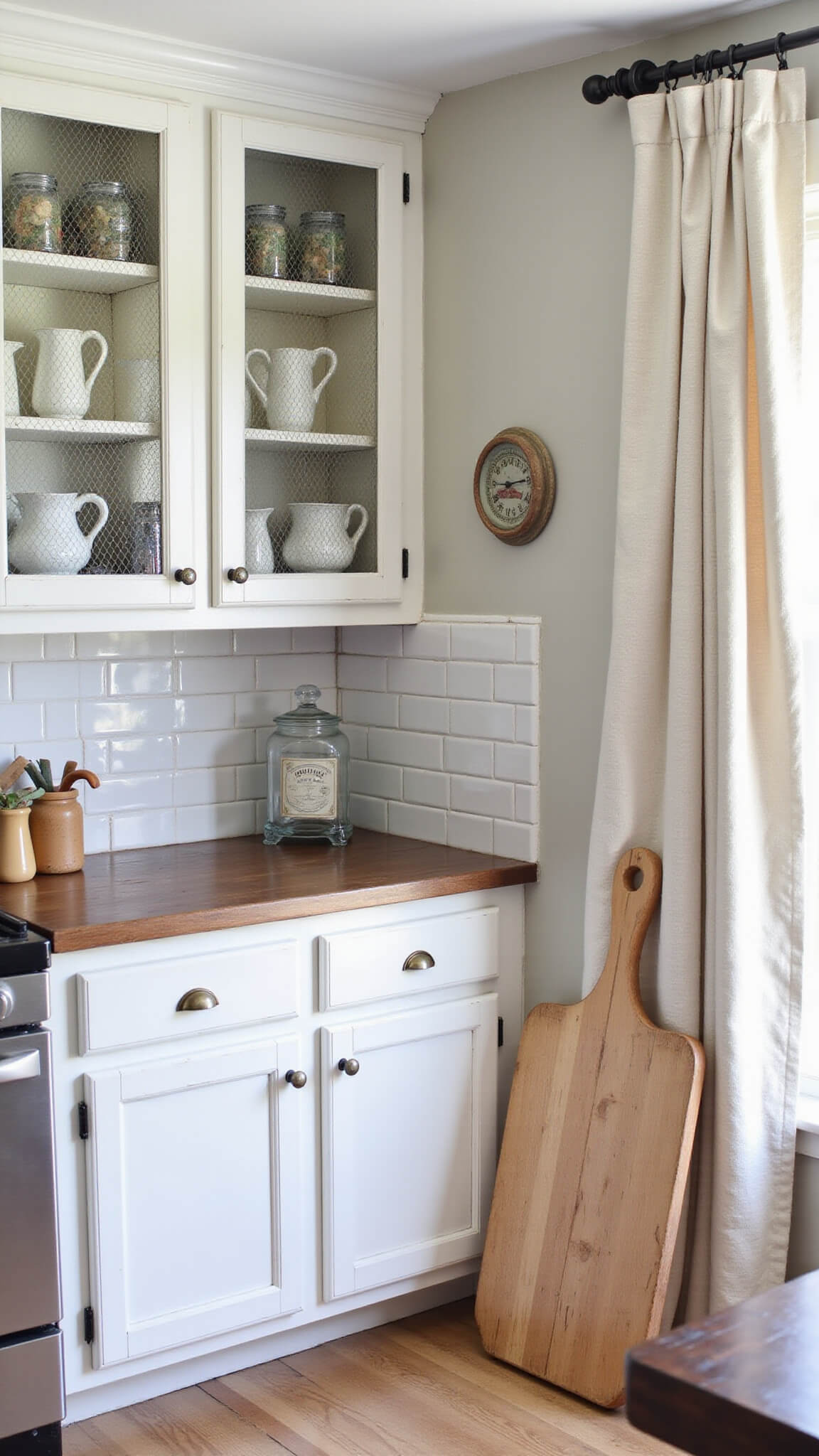 Cozy farmhouse kitchen corner with white distressed cabinets, chicken wire inserts, open shelves holding ironstone pitchers and mason jars, and an antique bread board against a subway tile backsplash in soft natural light.