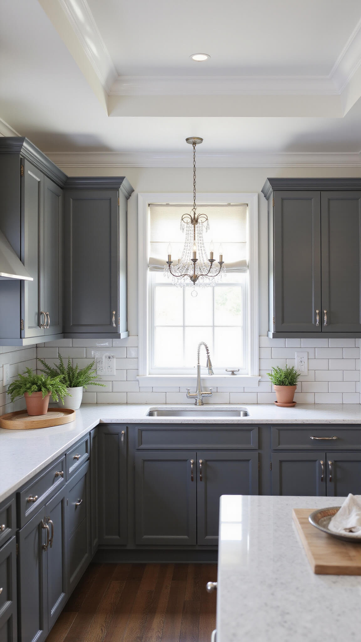 Transitional 16x14ft kitchen with tray ceiling, charcoal gray cabinets, quartz counters, crystal chandelier, white subway tile backsplash, and soft daylight filtering through roman shades.