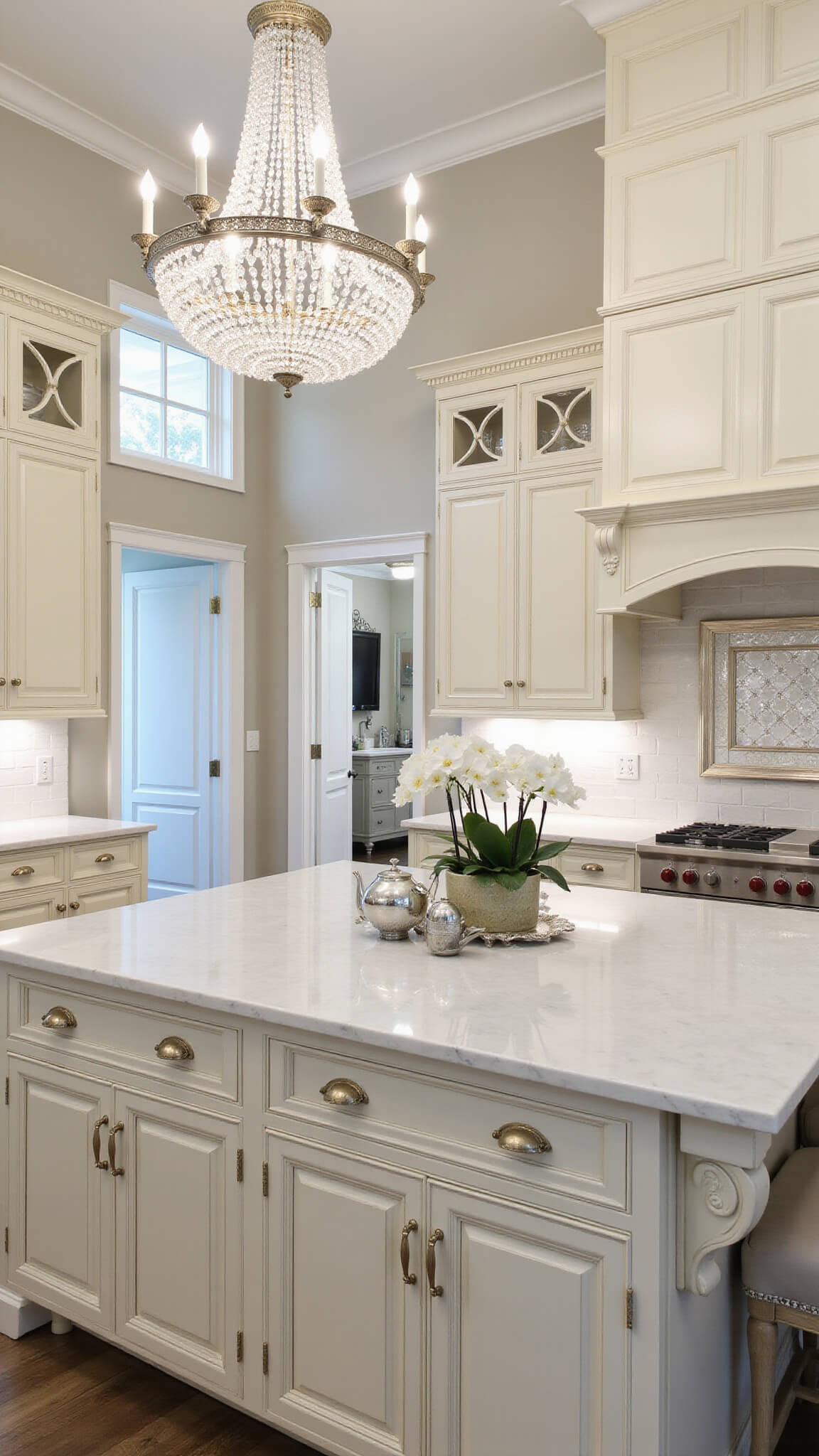 Elegant double-height cream kitchen with crystal chandelier, marble surfaces, antique mirror backsplash, and silver accents in soft morning light.