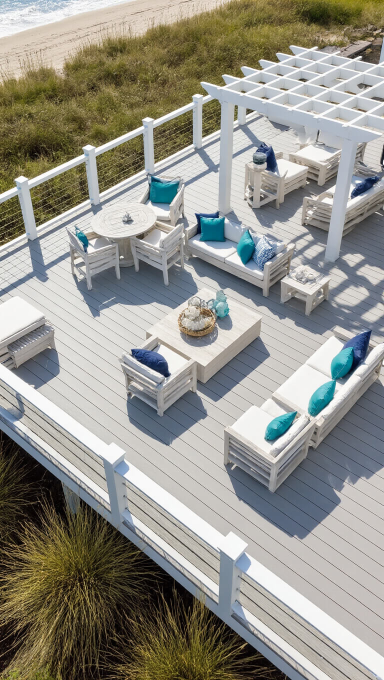 Overhead drone view of L-shaped driftwood gray deck with whitewashed furniture, navy and aqua pillows, glass railings, coastal grasses, and a pergola casting dappled shade.
