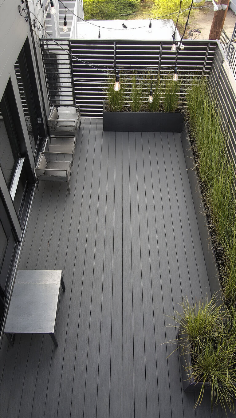 Overhead view of 16x18ft urban deck with dark metallic decking, steel railings, concrete and metal furniture, corrugated planters with tall grasses, and Edison bulb string lights under harsh direct lighting.