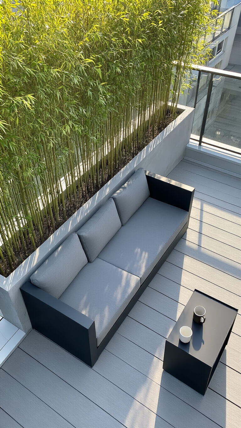 Modern minimalist balcony at dawn with grey loveseat, bamboo planters, black side table, and morning shadows on light grey decking.