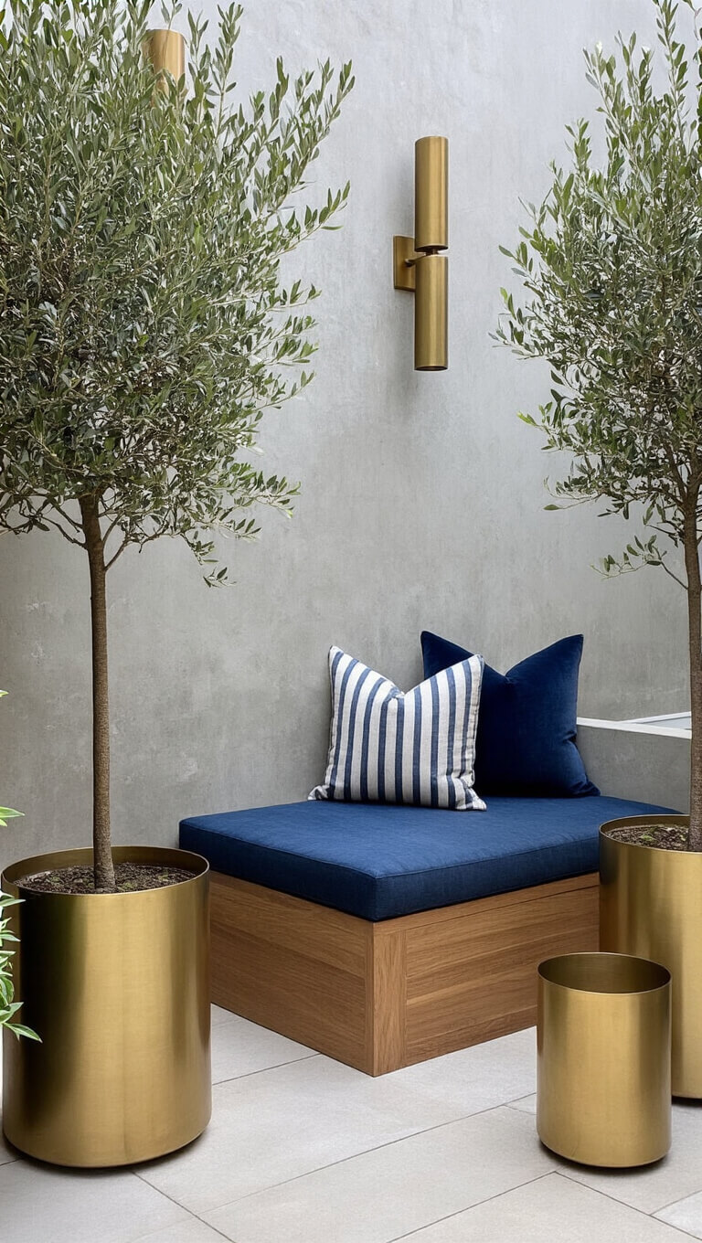 Corner view of luxe urban patio with navy cushioned teak bench, brass planters with olive trees, and copper sconces on textured concrete wall.