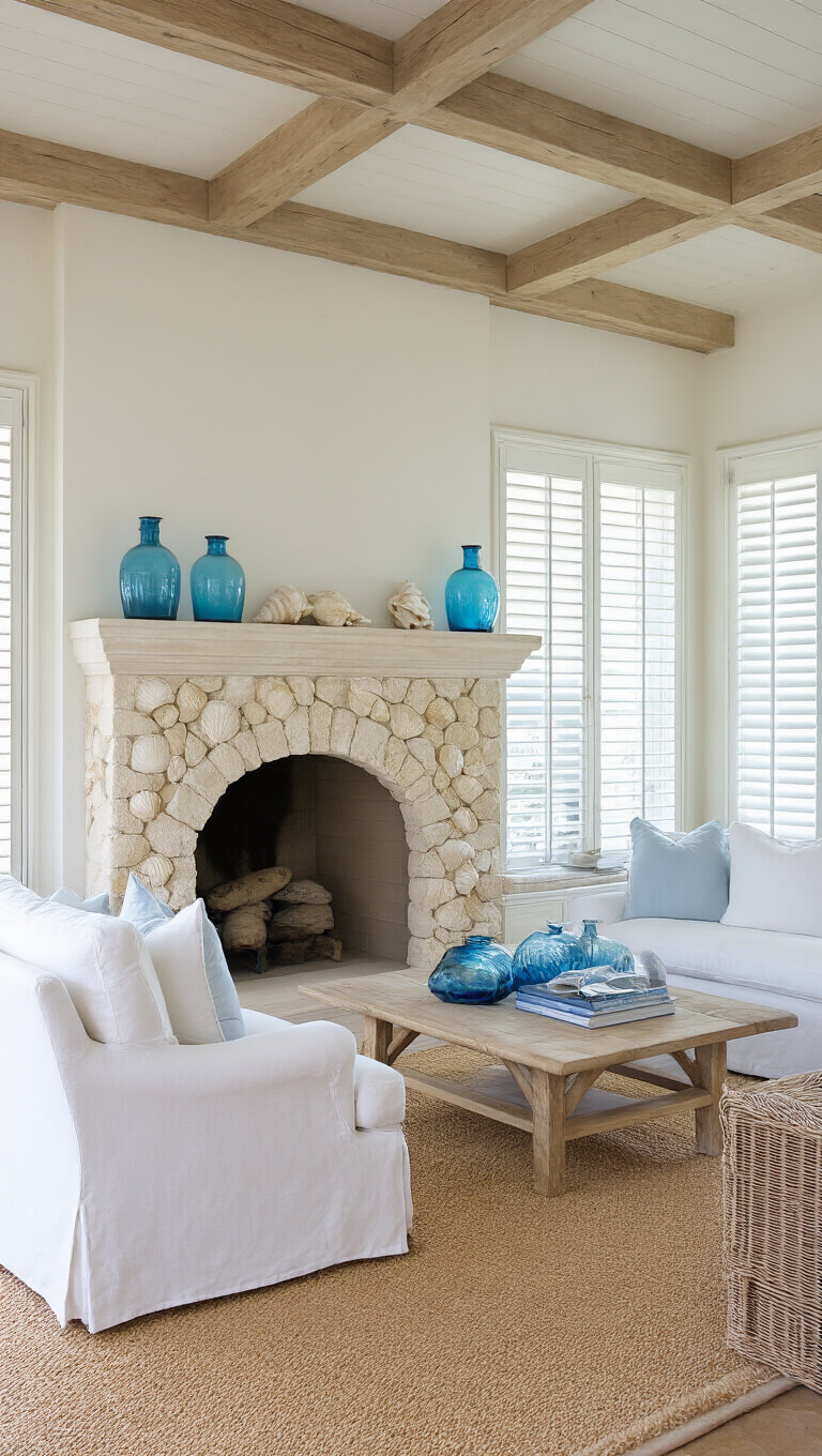 Bright coastal living room with white slipcovered furniture, sisal rug, beamed ceiling, limestone fireplace with shell stone surround, and morning light filtering through plantation shutters.
