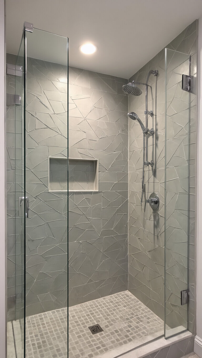 Walk-in shower with soft grey geometric tile feature wall, chrome fixtures, and frameless glass under warm evening lighting.